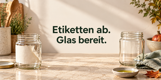 Ein Glas mit teilweise gelöstem Papieretikett neben einem sauberen Glas, Tuch und Ölschälchen; Titel: Etiketten ab. Glas bereit.